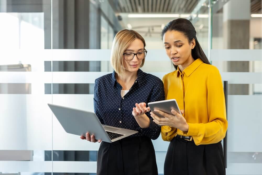 Two businesswomen standing in an office, discussing a project while holding a laptop and a digital tablet, illustrating teamwork, communication and modern technology in business