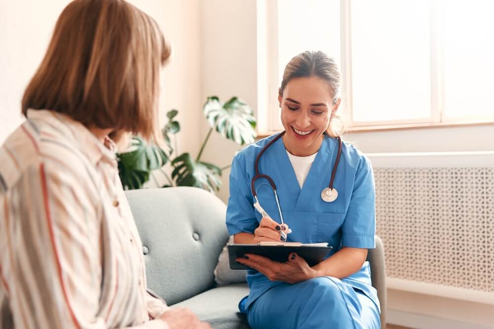 Happy young female nurse provide care medical service help support smiling mature woman at homecare medical visit, lady carer doctor give empathy encourage retired patient sit on sofa at home hospital