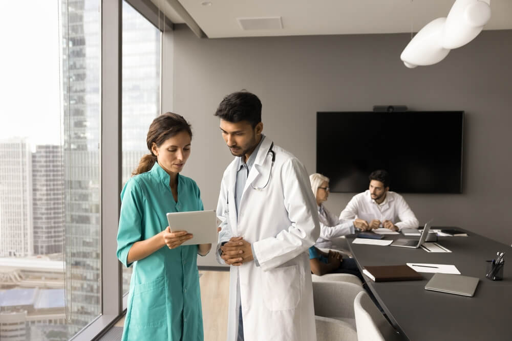 Serious female doctor consulting male colleague on corporate meeting in clinic conference room, showing tablet to boss, speaking, asking for advice. Two medical professionals using healthcare app