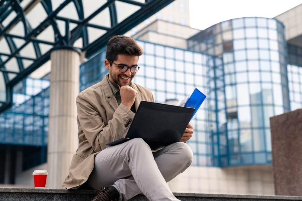 A joyful young Caucasian male entrepreneur reviews paperwork and utilizes a laptop while seated on steps outside an office building, wearing casual business attire, with a coffee cup nearby.