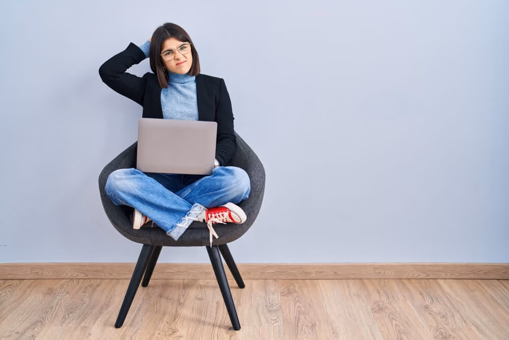 Young hispanic woman sitting on chair using computer laptop confuse and wondering about question. uncertain with doubt, thinking with hand on head. pensive concept.