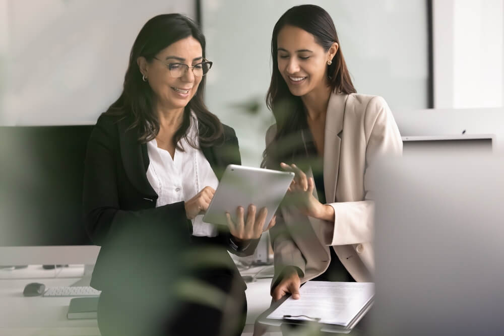 Positive younger Maori business professional girl showing new work application on tablet to mature colleague woman, touching screen of gadget, smiling. Through object candid blurred view