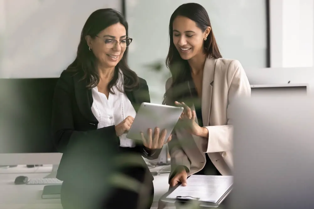 Positive younger Maori business professional girl showing new work application on tablet to mature colleague woman, touching screen of gadget, smiling. Through object candid blurred view