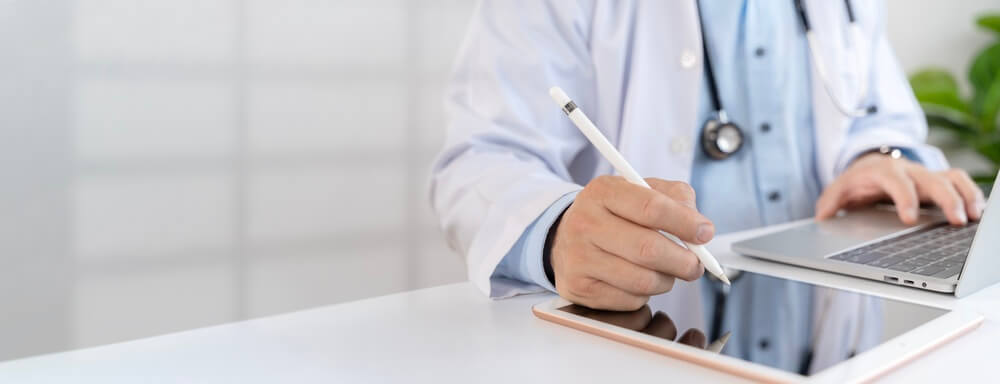 Cropped doctor using stylus on tablet beside laptop in a clean clinic office, ideal for telemedicine, digital healthcare, patient records and medical marketing with copy space.