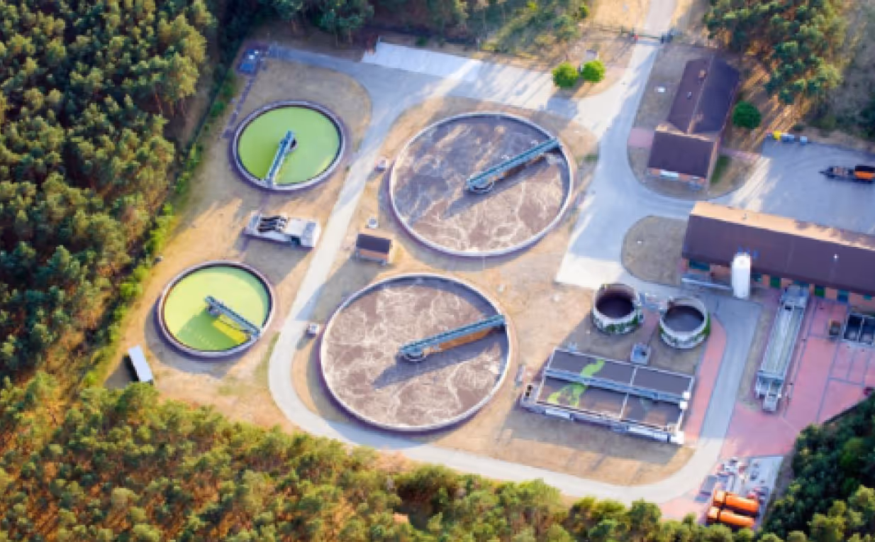Aerial view of a wastewater treatment plant with circular clarifiers and surrounding forest.