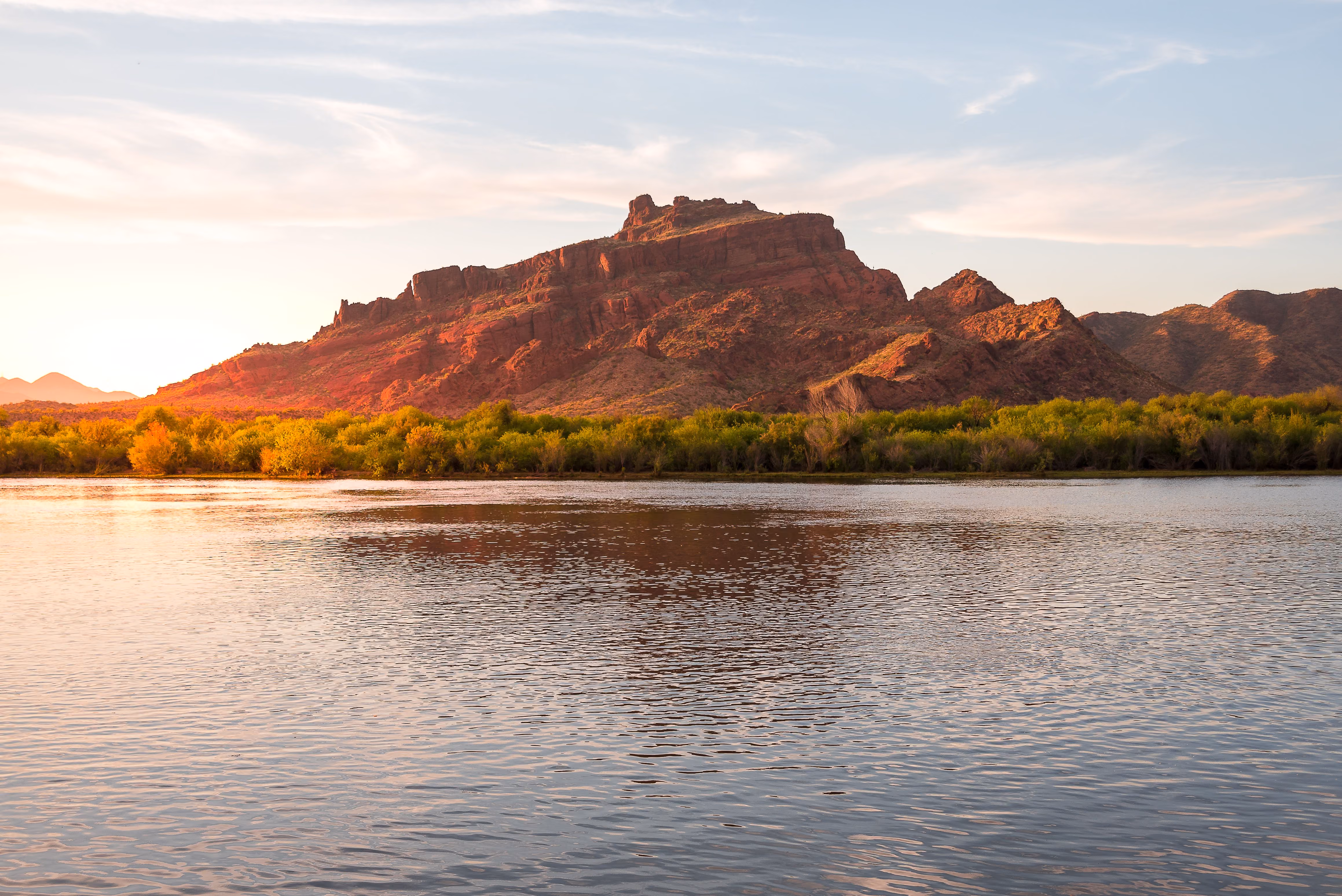 Calm river in front of a reddish rocky mountain with green shrubs along the riverbank at sunset under a partly cloudy sky.