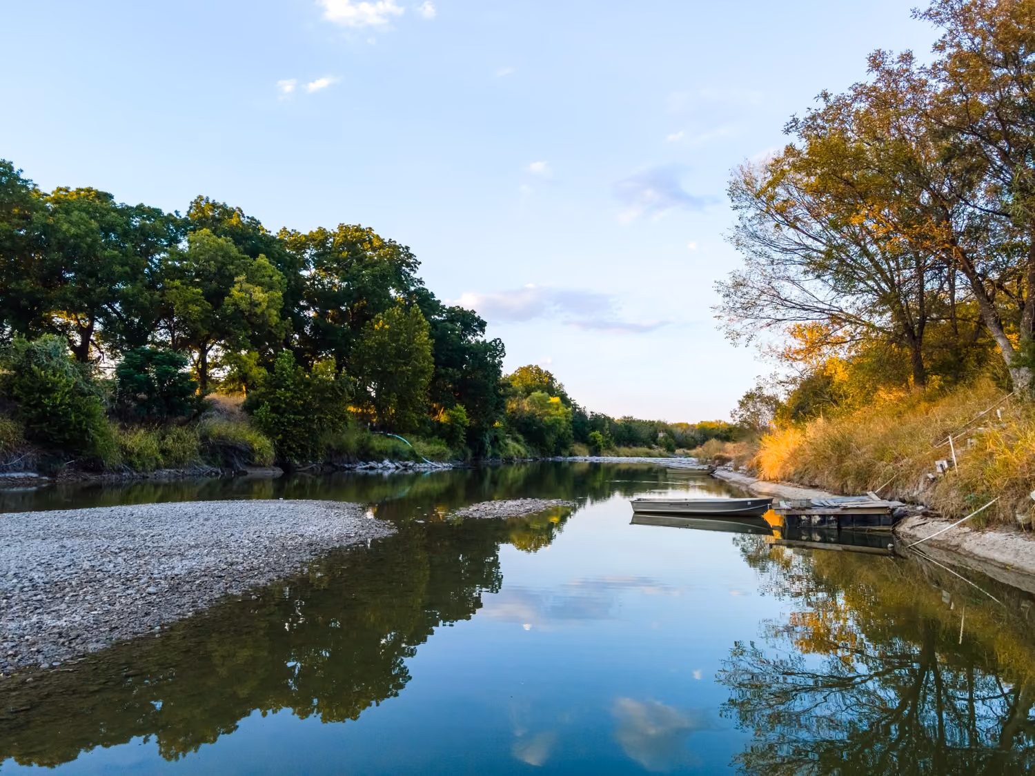 Calm river with clear reflections of trees and sky, bordered by green trees on the left and sparse trees with dry grass on the right, with a small dock or boats tied along the right bank.