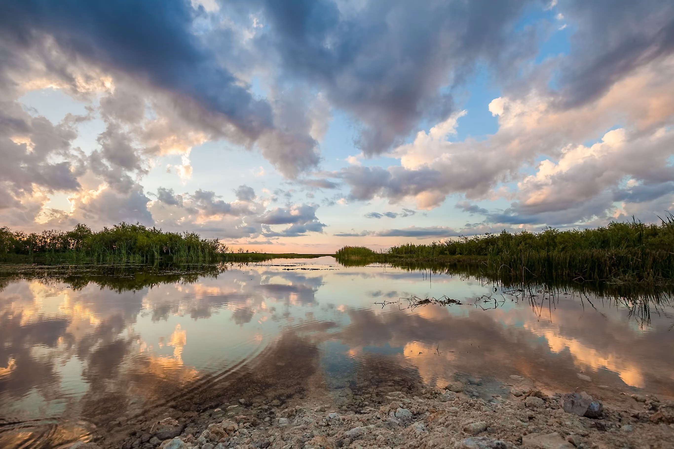 Calm water body reflecting a dramatic sky with clouds at sunset, bordered by green vegetation and rocky shore in the foreground.