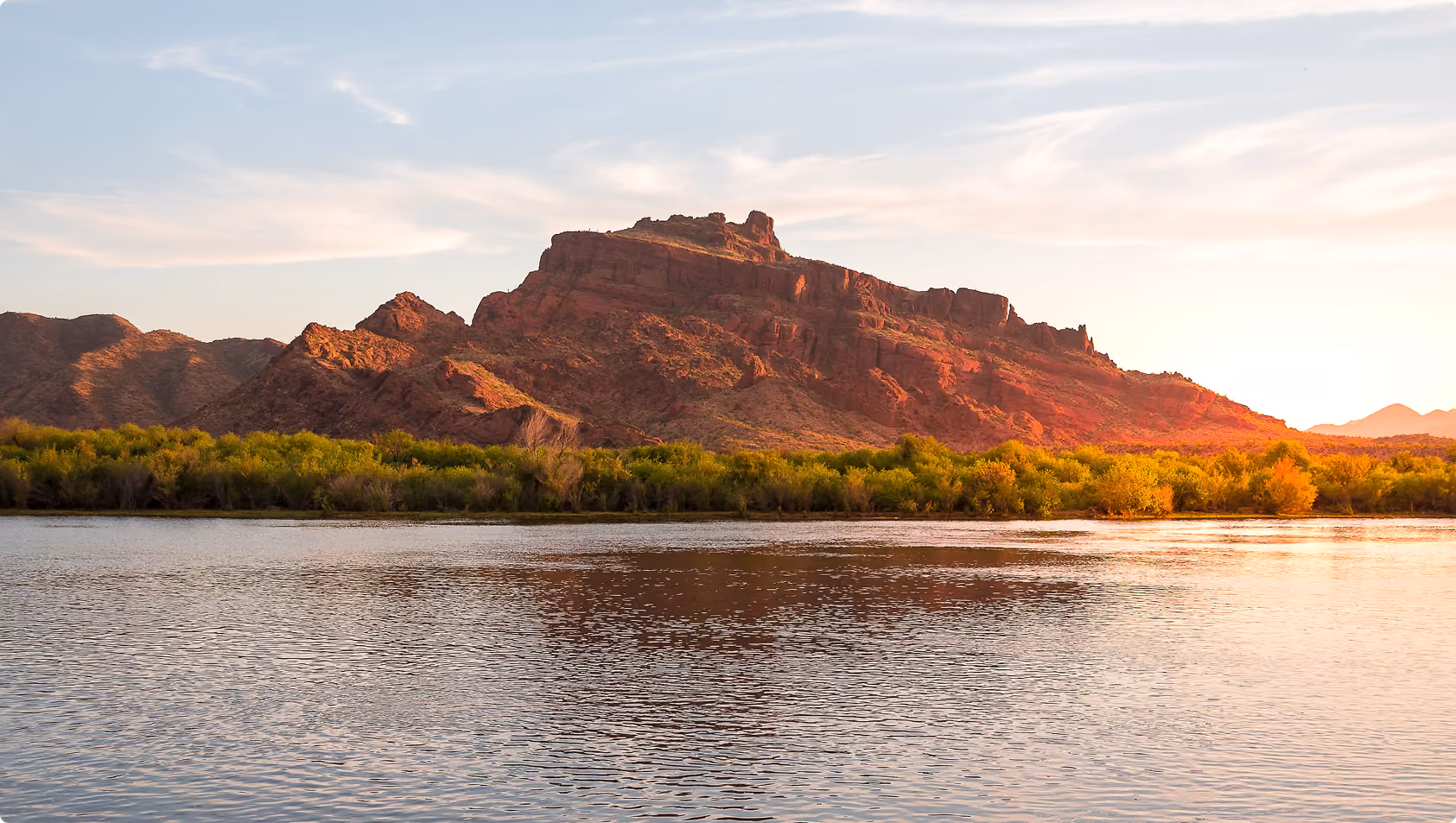 Red rocky mountain bathed in warm sunlight with green trees at its base and a calm reflective river in the foreground under a partly cloudy sky.