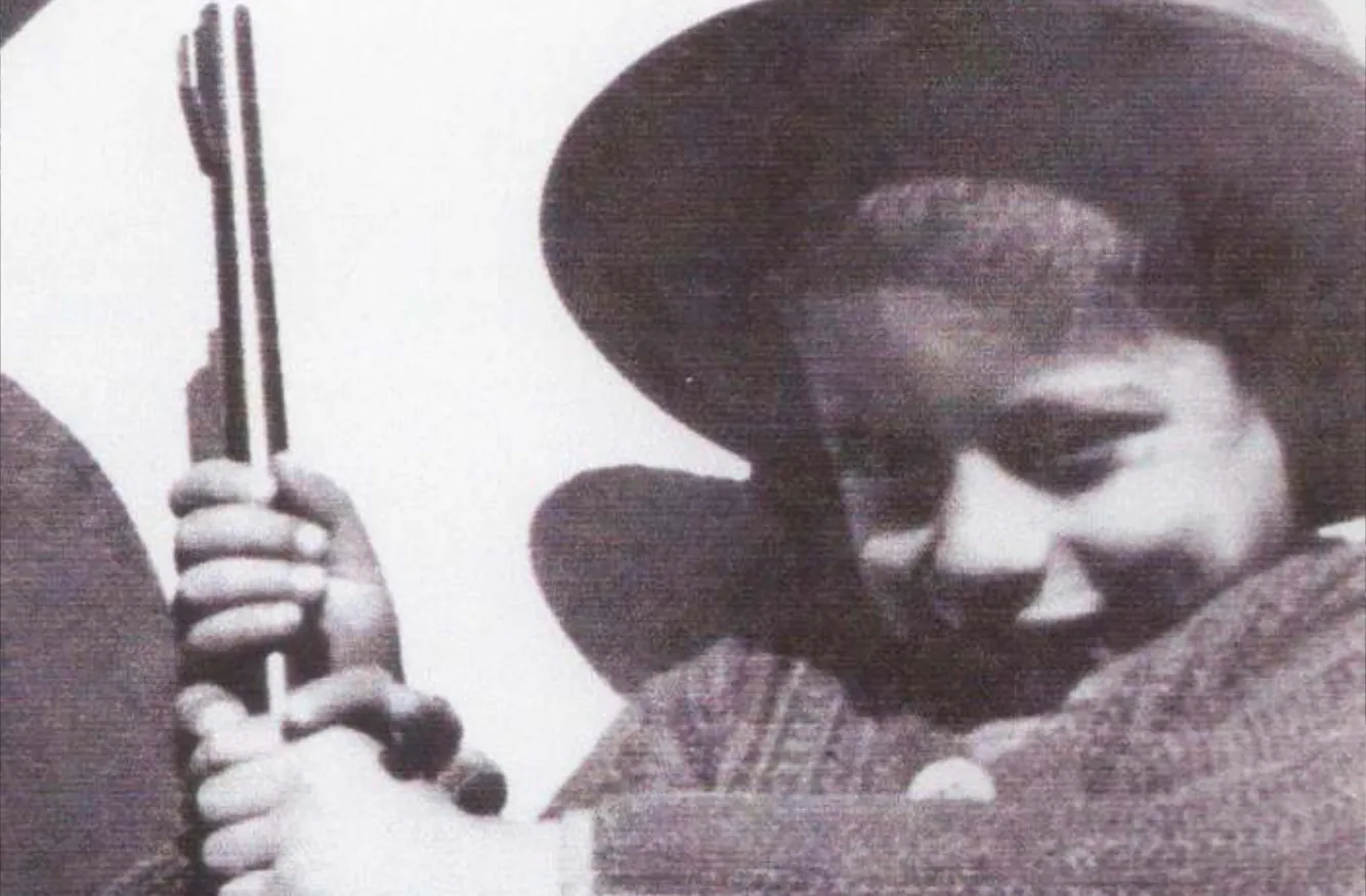 Black and white close-up of a smiling young boy wearing a hat and holding stringed musical instrument.