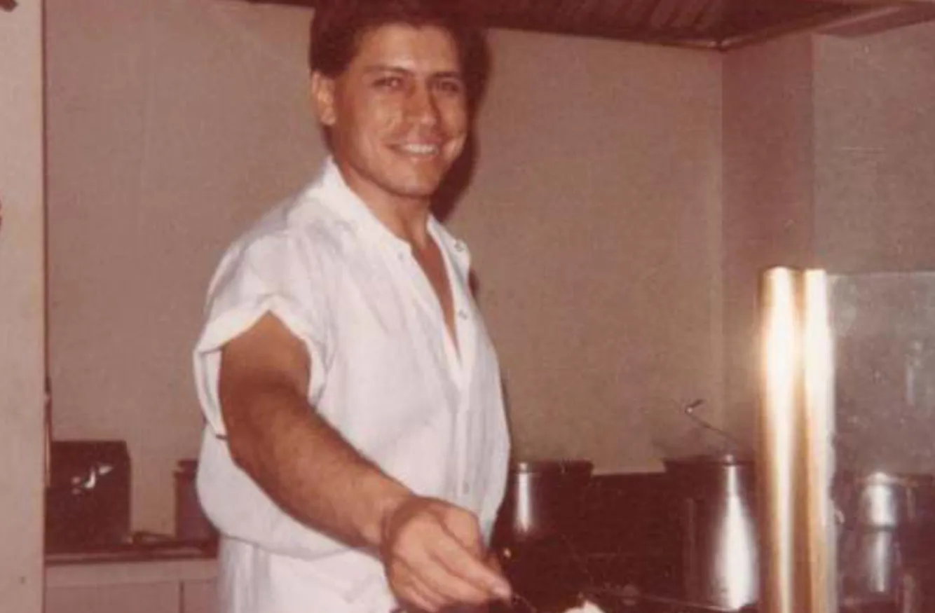 Smiling man in a white shirt stands in a kitchen holding a utensil, with cooking pots in the background.