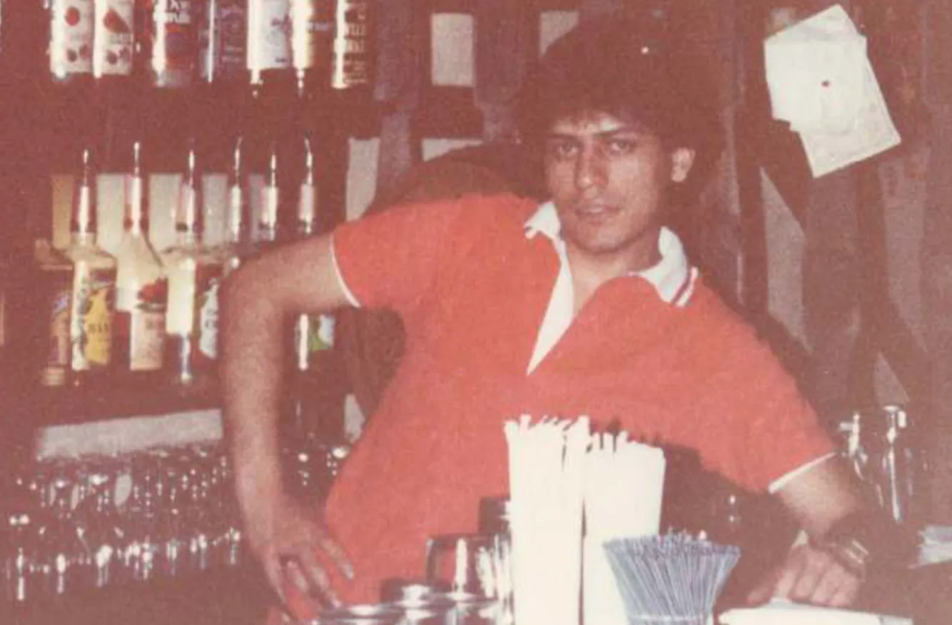 Man in a red polo shirt standing behind a bar counter with bottles and glasses in the background.