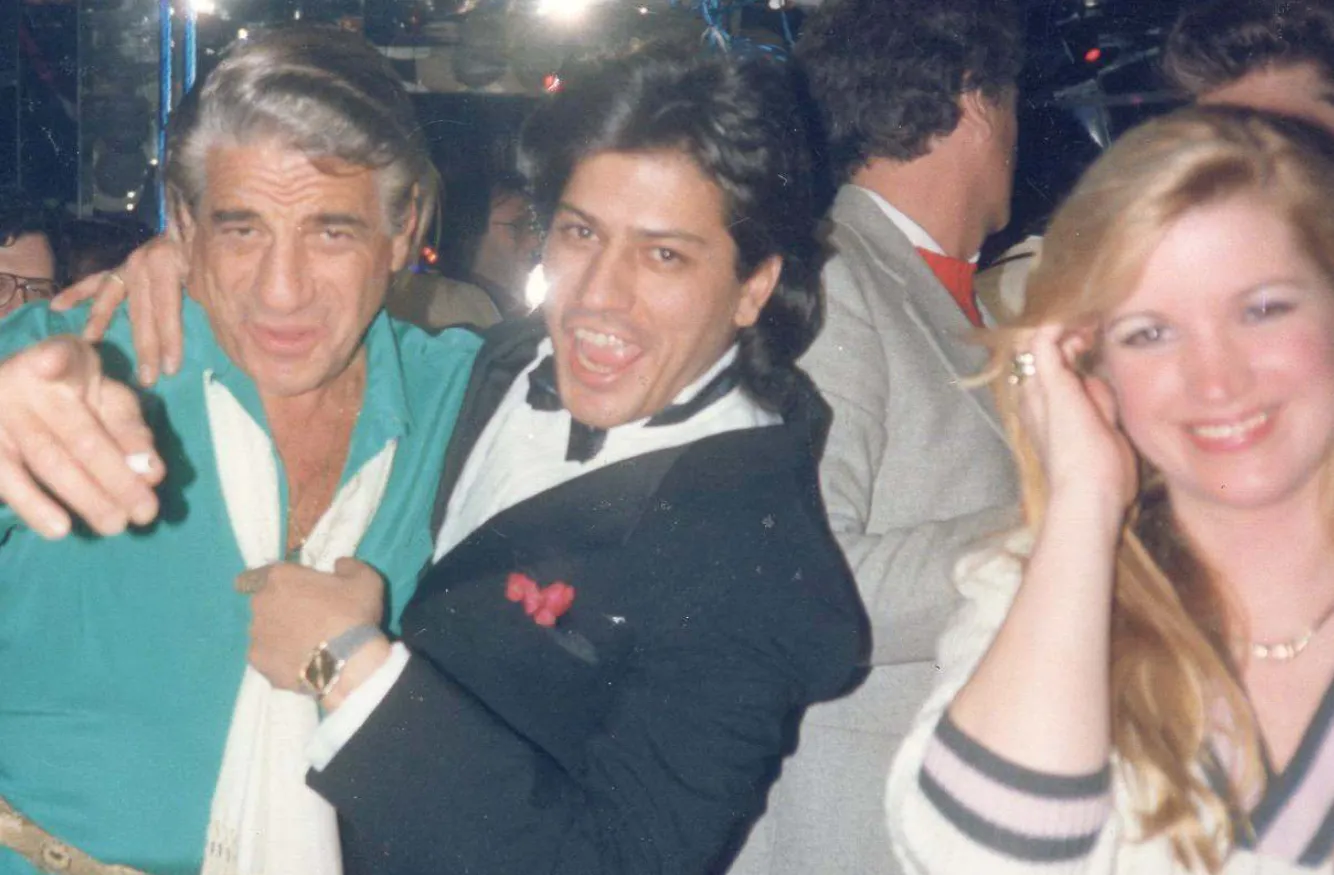 Two men posing energetically together at a party, one in a teal shirt and the other in a black tuxedo, with a smiling woman in a white sweater on the right.