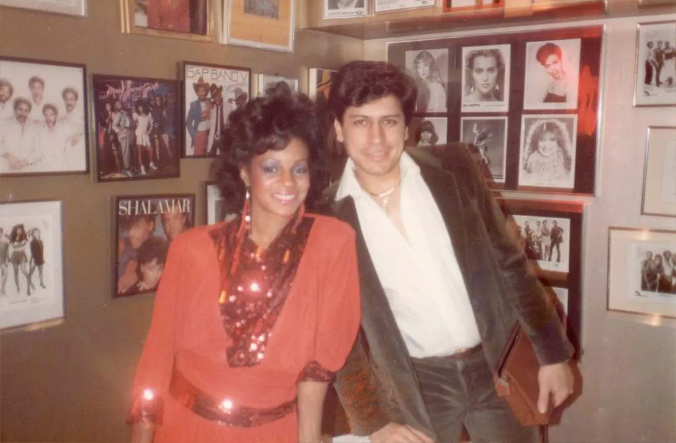 A woman in a red sequined dress posing with a man in a white shirt and dark jacket in front of a wall decorated with music and celebrity photos.