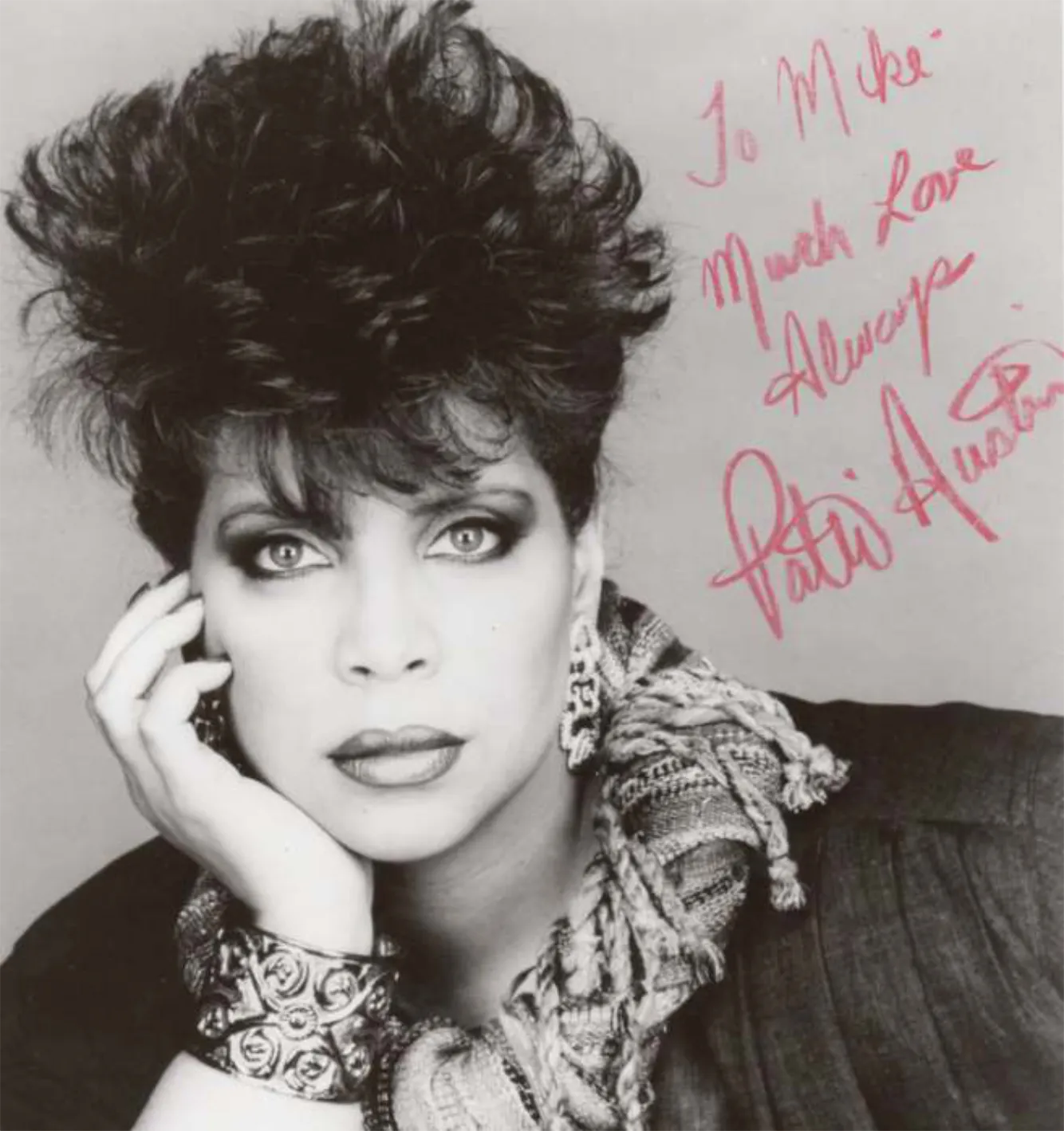 Black and white portrait of a woman with voluminous curly hair, wearing a patterned scarf, large earrings, and a decorative wrist cuff, resting her face on her hand with an autograph written in red ink on the right side.