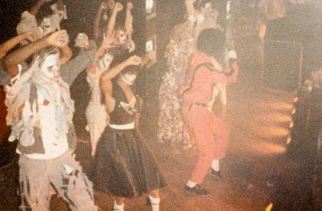 A group of people dressed in costumes, some with white face paint, dancing with their arms raised in a dimly lit room.