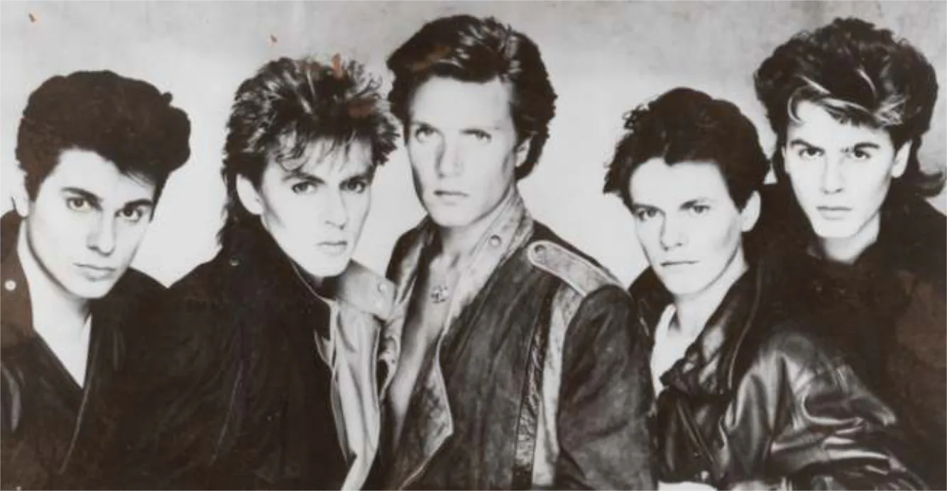Black-and-white photo of five men with 1980s hairstyles and jackets, posed closely together against a plain background.