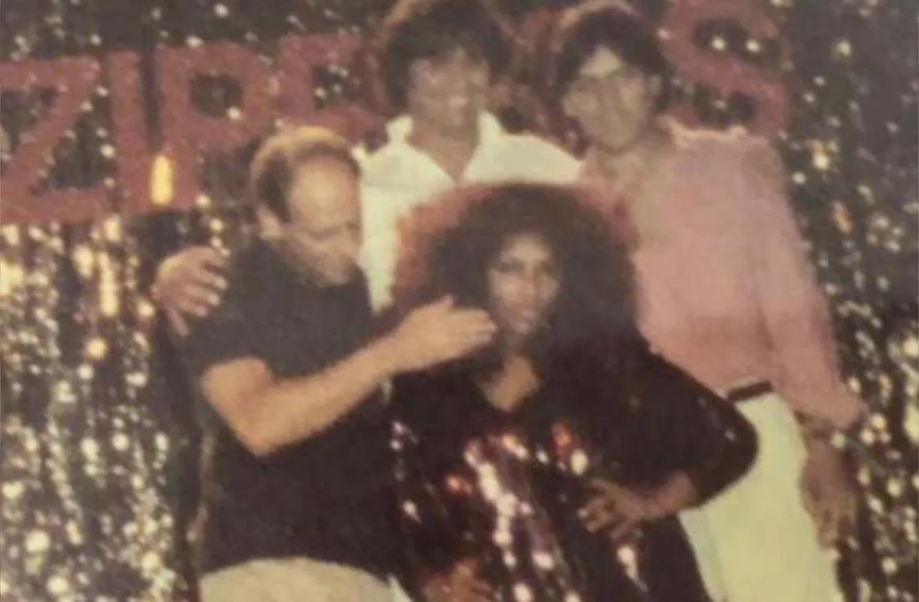 A group of four people posing together in front of a backdrop with large letters and glittery decorations, with a woman in front wearing a shiny dark outfit and three men standing behind her.