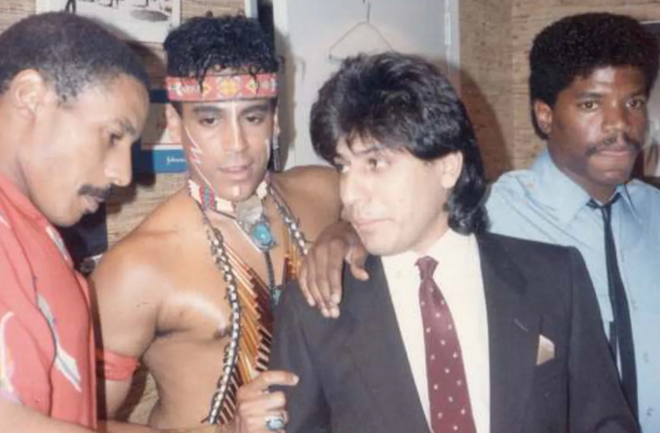 Four men standing closely together, one dressed in tribal-inspired costume with painted face and beaded accessories, another in a suit and tie, and two others in shirts, one looking down and the other looking forward.