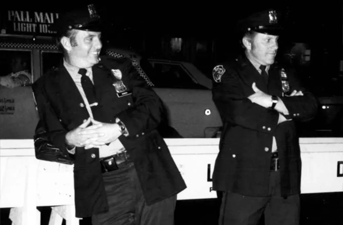 Two police officers in uniform standing and leaning against a barricade at night, one smiling and the other with arms crossed.