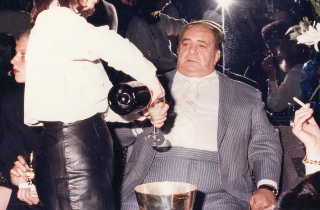 A man in a pinstripe suit sitting as a woman pours champagne into his glass at a dimly lit social gathering.