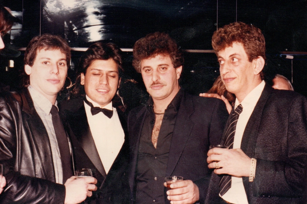 Four men dressed formally, three holding drinks and standing closely together in a social setting.