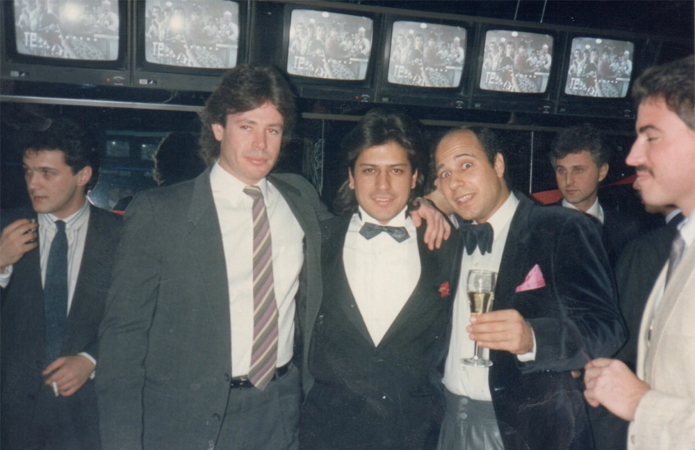 Group of six men in formal attire, including suits and tuxedos, standing together indoors with several small TV screens in the background.