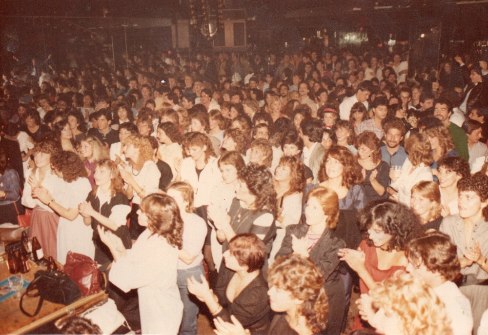 Large crowd of people standing closely together in a dimly lit indoor venue, many clapping and facing forward.