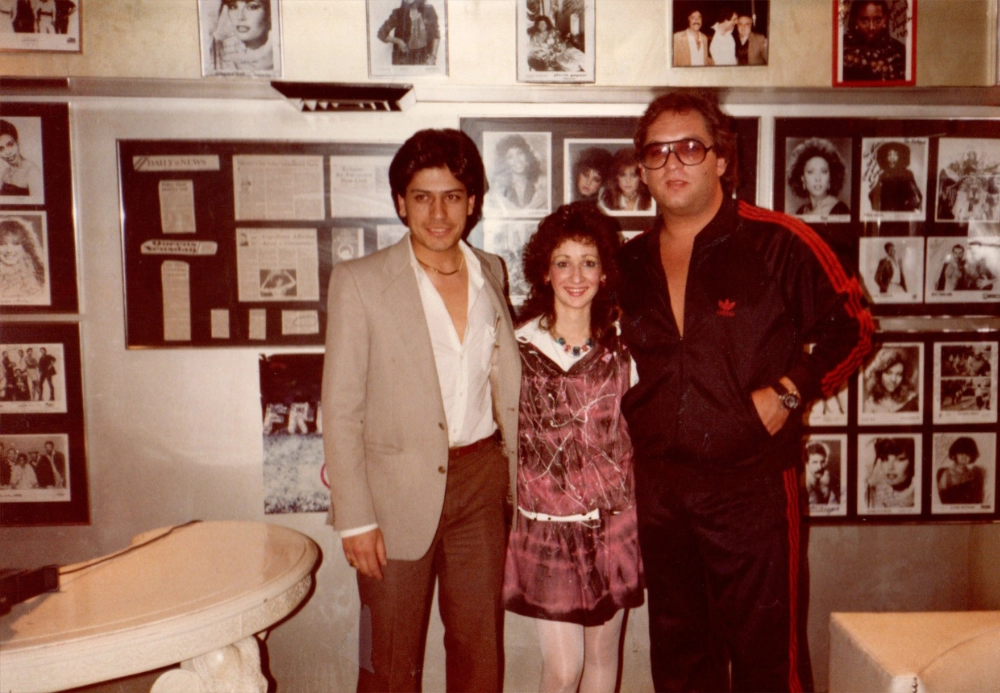 Three people posing indoors in front of a wall covered with framed photographs and newspaper clippings; the woman in the center wears a pink dress, the man on the left wears a beige blazer and brown pants, and the man on the right wears a black tracksuit with red stripes and large sunglasses.