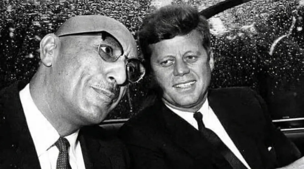 Black and white photo of two men in suits sitting closely together inside a car, smiling.