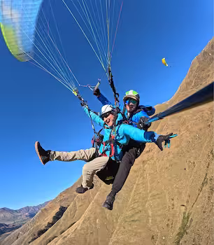 Two people tandem paragliding over a dry mountainous landscape under a clear blue sky.