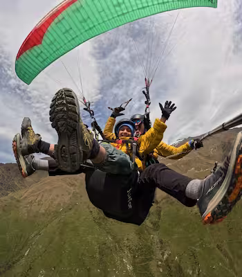 Two people tandem paragliding over a green mountainous landscape under cloudy sky.