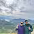 Two people standing together outdoors with a scenic valley and cloudy sky in the background.