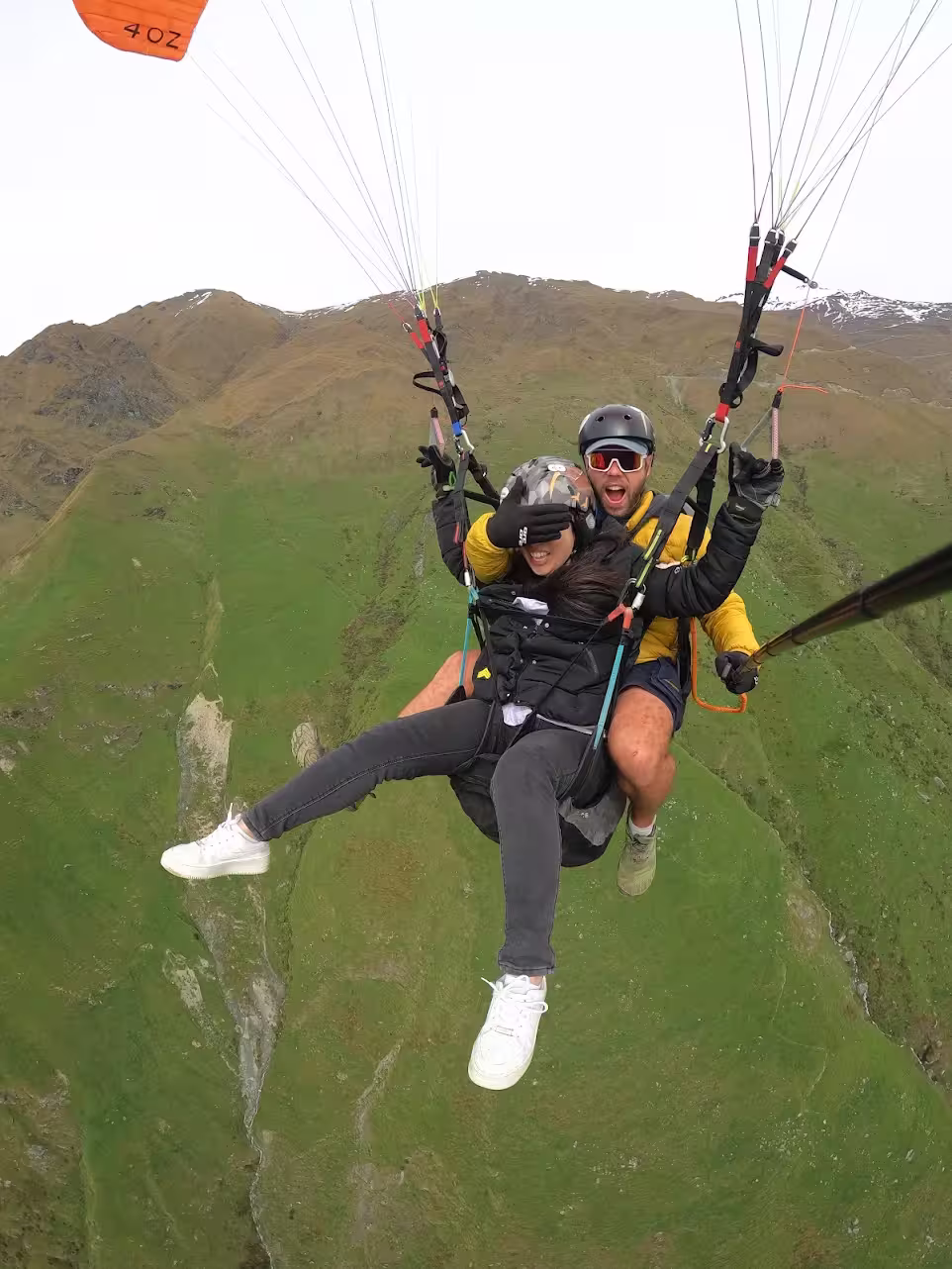 Two people in helmets and winter gear giving thumbs up, standing on snowy ground with paragliding equipment behind them.
