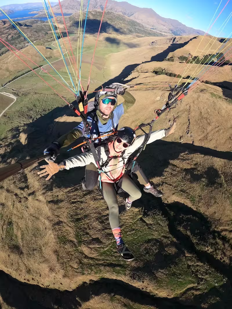 Two people tandem paragliding over brown and green hilly terrain under a clear blue sky.
