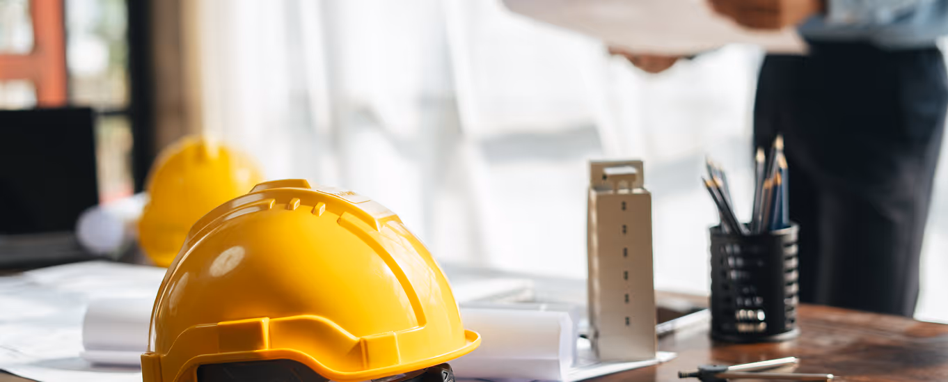 Yellow construction helmets on a table with architectural blueprints and pens, a person shaking hands in the background.