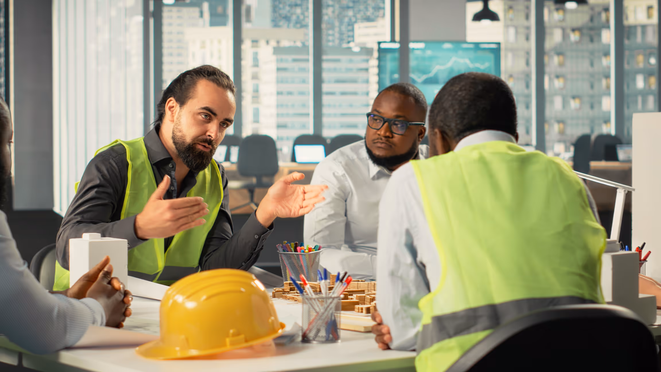Three construction workers in safety vests discussing a project at a table with a model and helmet.