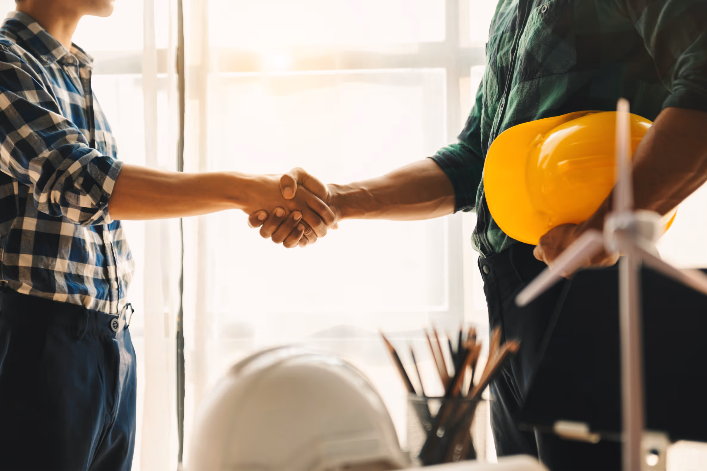 Two workers shaking hands indoors, one holding a yellow construction helmet in a bright room.