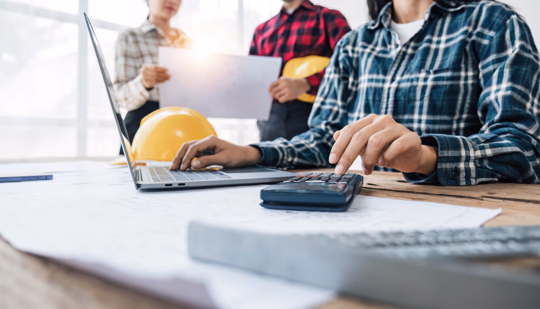 Person in plaid shirt working on a calculator with laptop and hard hats nearby on the desk.