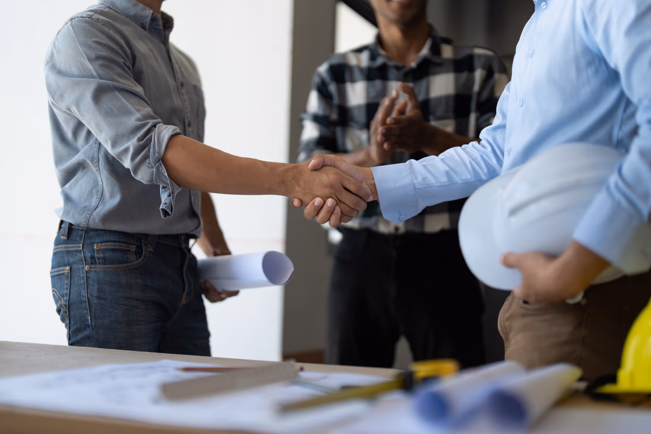 Two men shaking hands in a meeting, one holding a white hard hat and the other holding rolled papers.