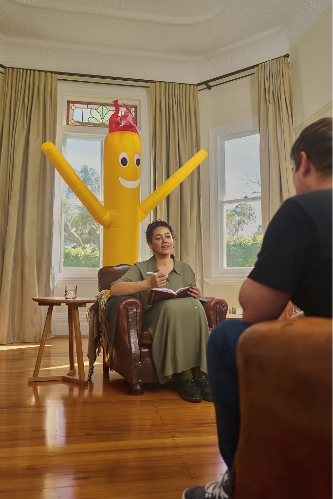Counsellor in an armchair taking notes with a client as a yellow inflatable tube man stands behind them