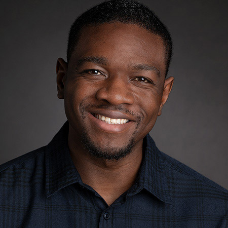 Smiling man with short hair and beard wearing a dark button-up shirt against a gray background.