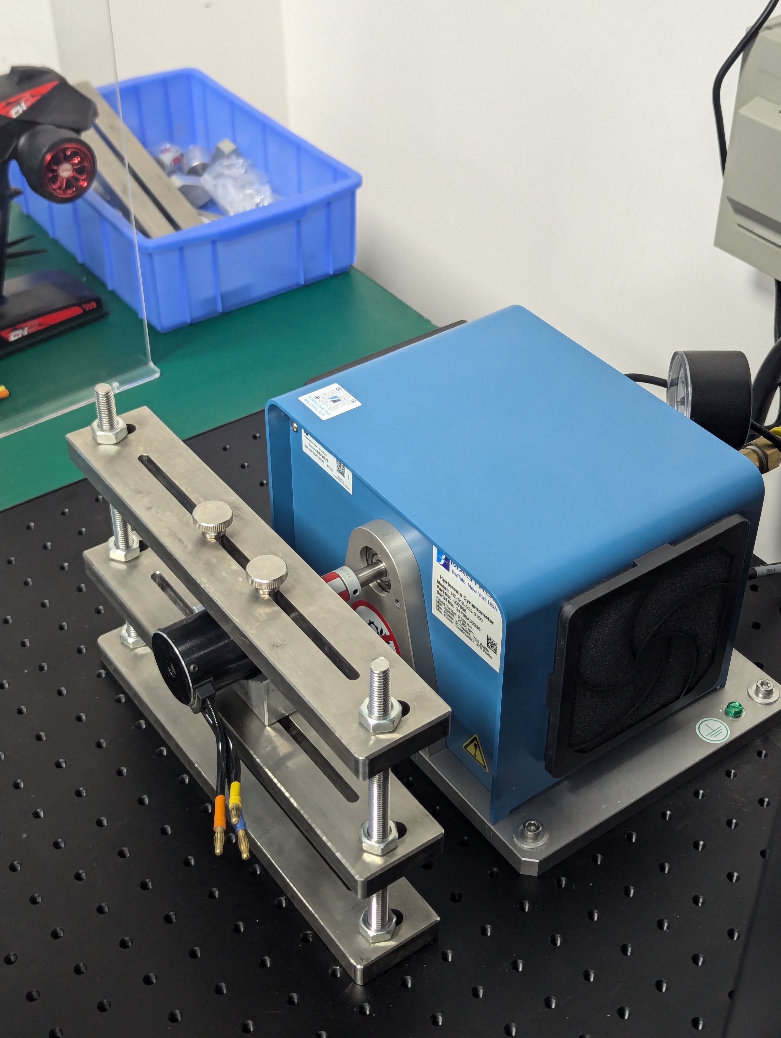 Blue laboratory pump device mounted on a black perforated metal surface with a metal clamp fixture and wires attached.