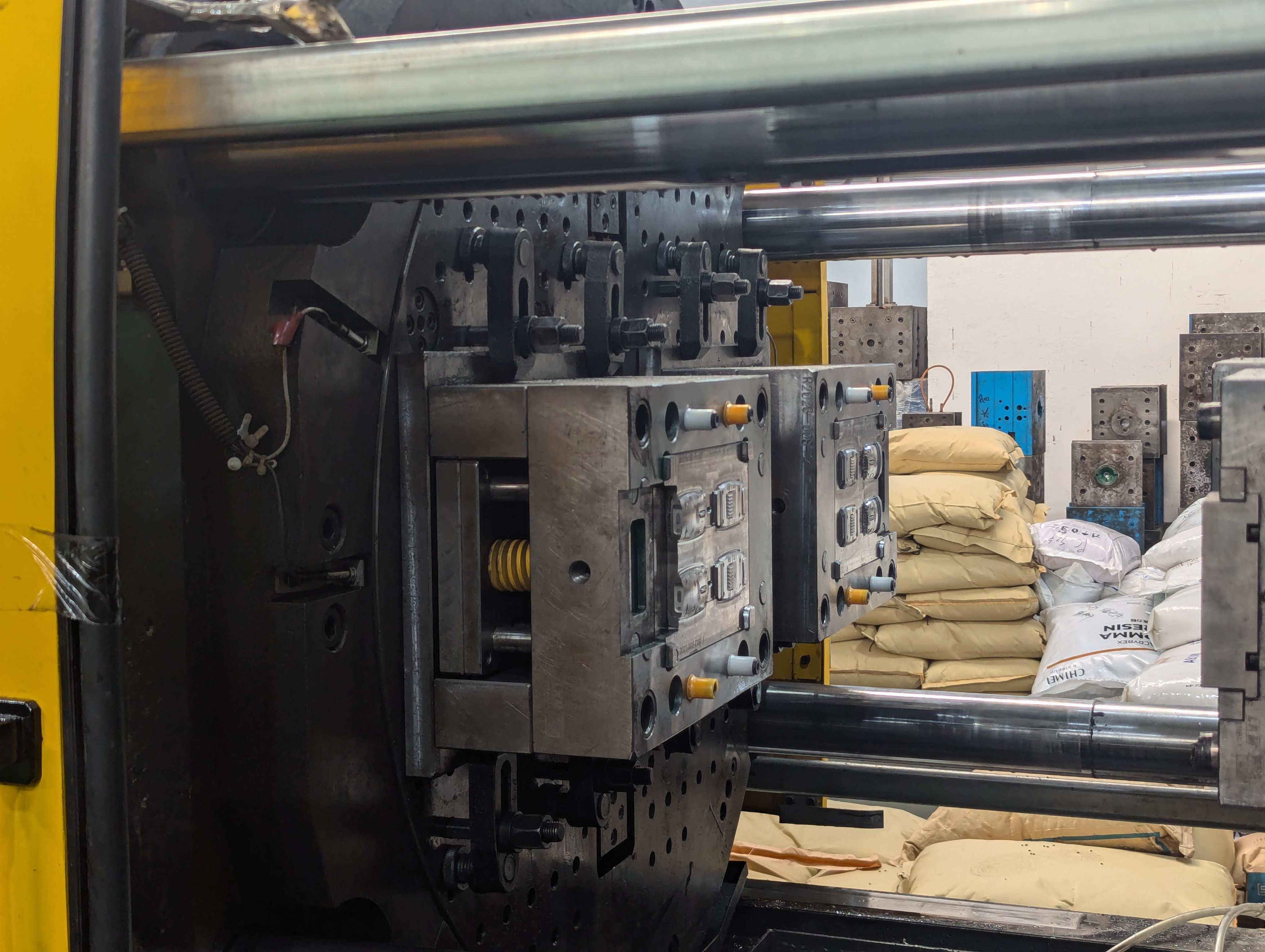 Close-up of an industrial injection molding machine with metal mold plates and stacked bags of raw material in the background.