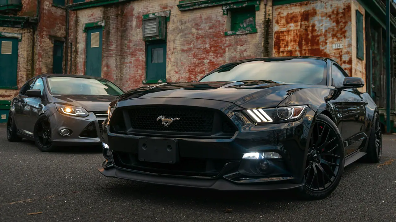 Zwei Autos geparkt vor einer alten Backsteinwand, vorn ein schwarzer Ford Mustang.