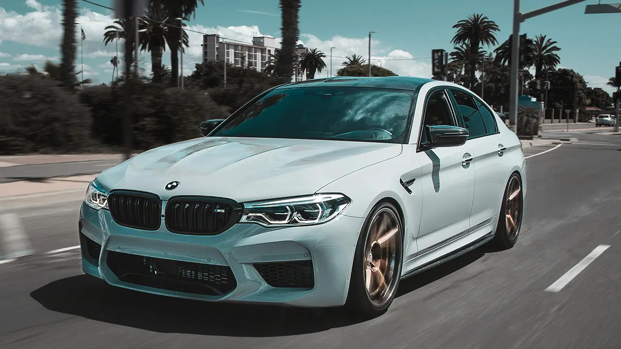 Weiße BMW M5 Limousine fährt auf einer Stadtstraße mit Palmen im Hintergrund.