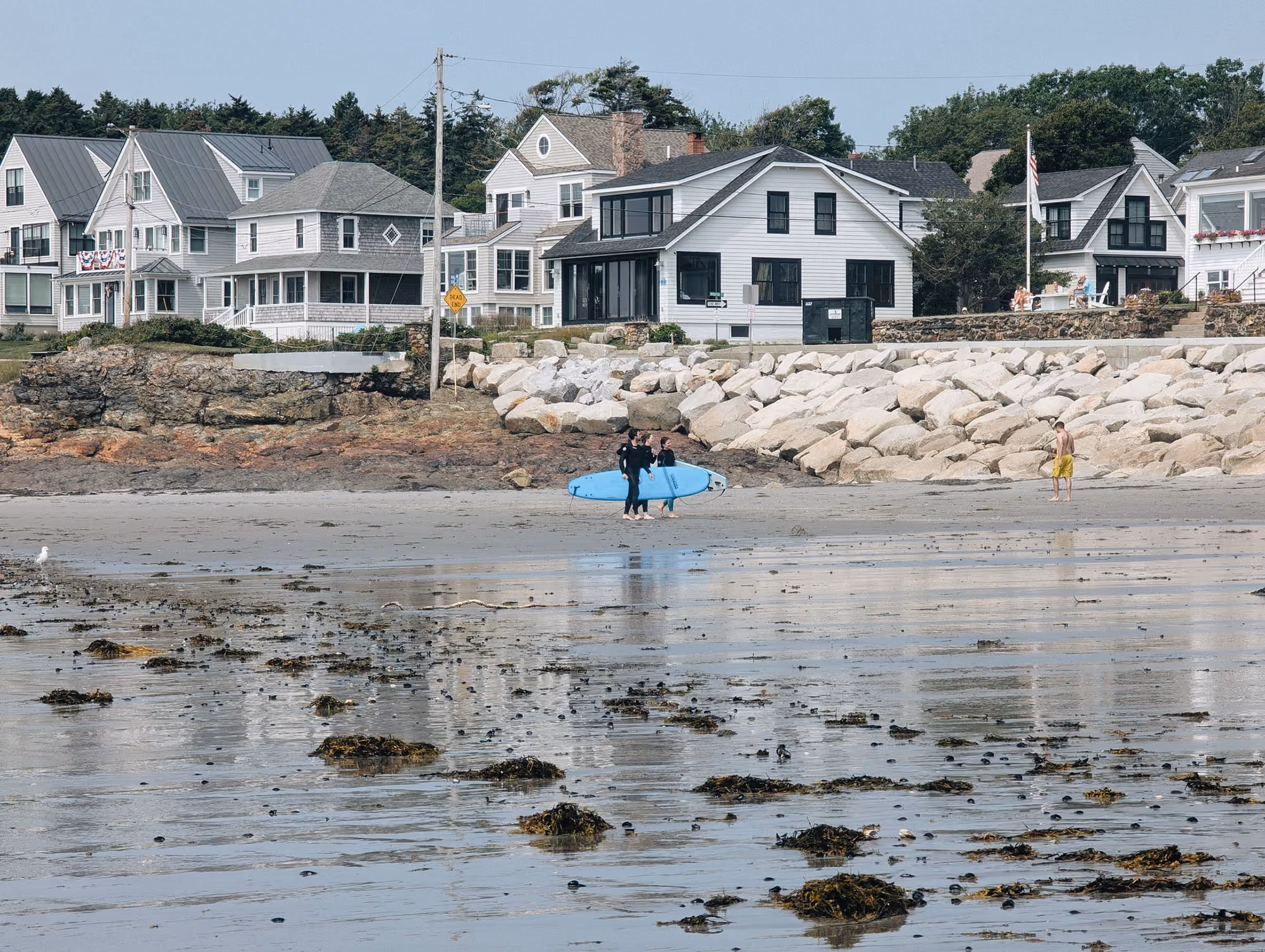 Three people with a blue surfboard on a sandy beach near rocky shoreline and waterfront houses.