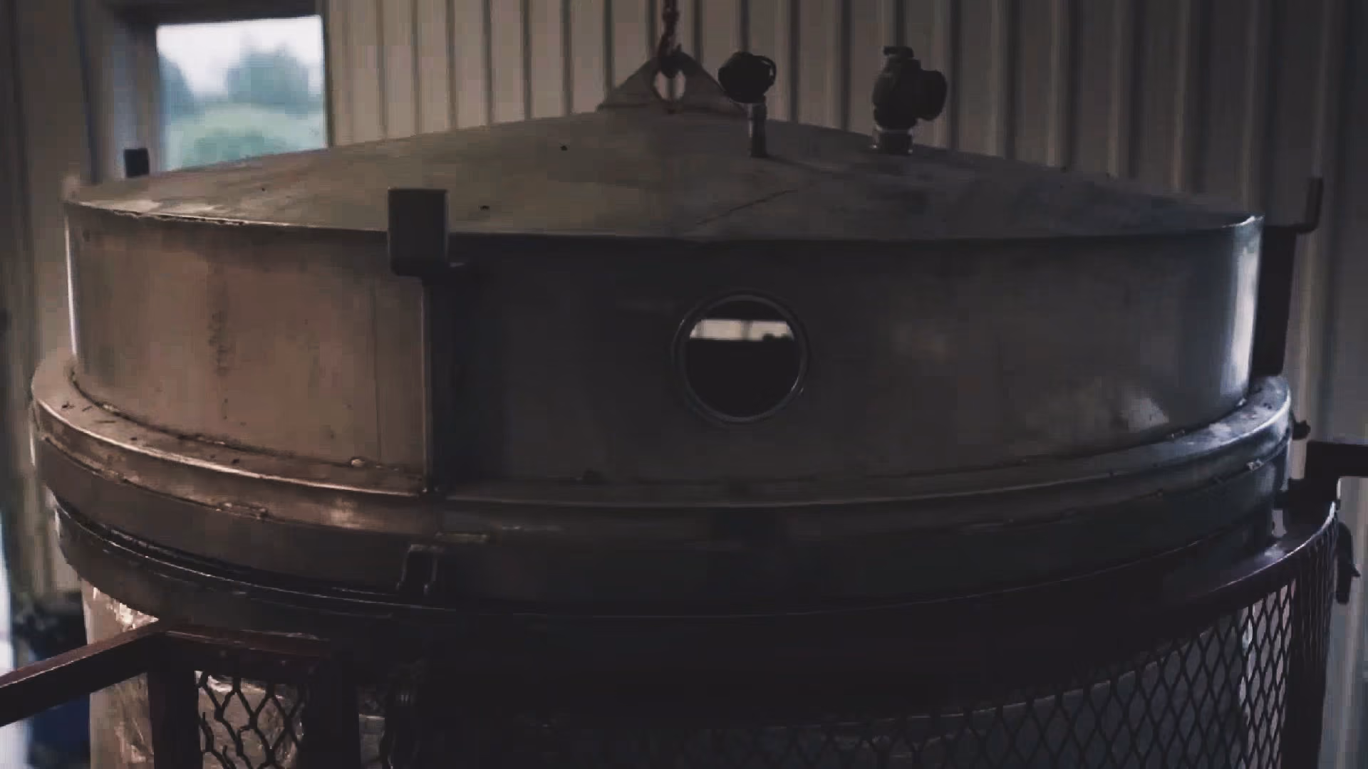 Large distiller metal tank with valves and a circular window in a dimly lit room.