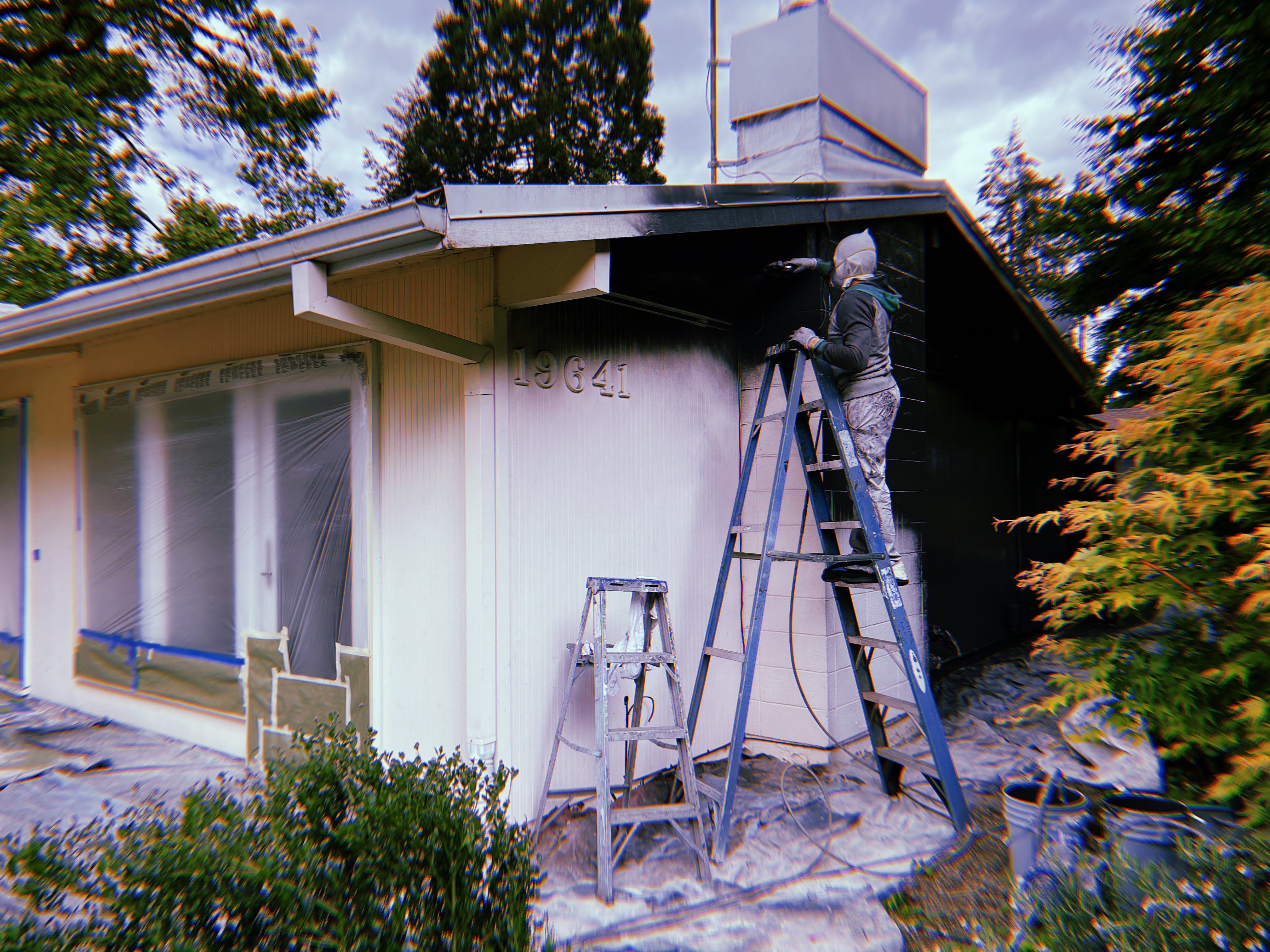 D'S Painting LLC painter on ladder applying exterior paint to a mid-century home with windows masked off