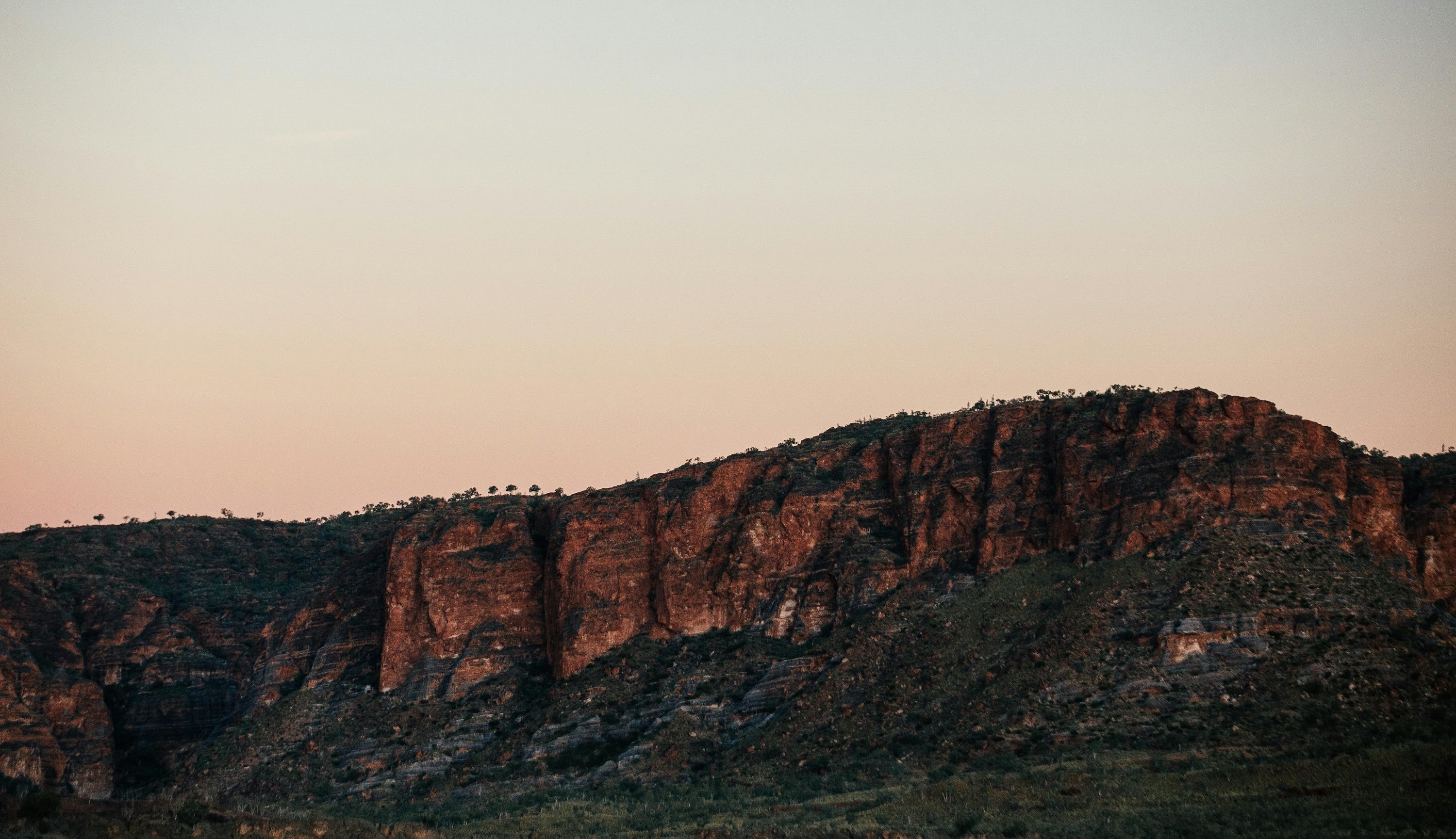 Rocky hills with reddish cliffs and sparse vegetation under a clear sky at sunset.